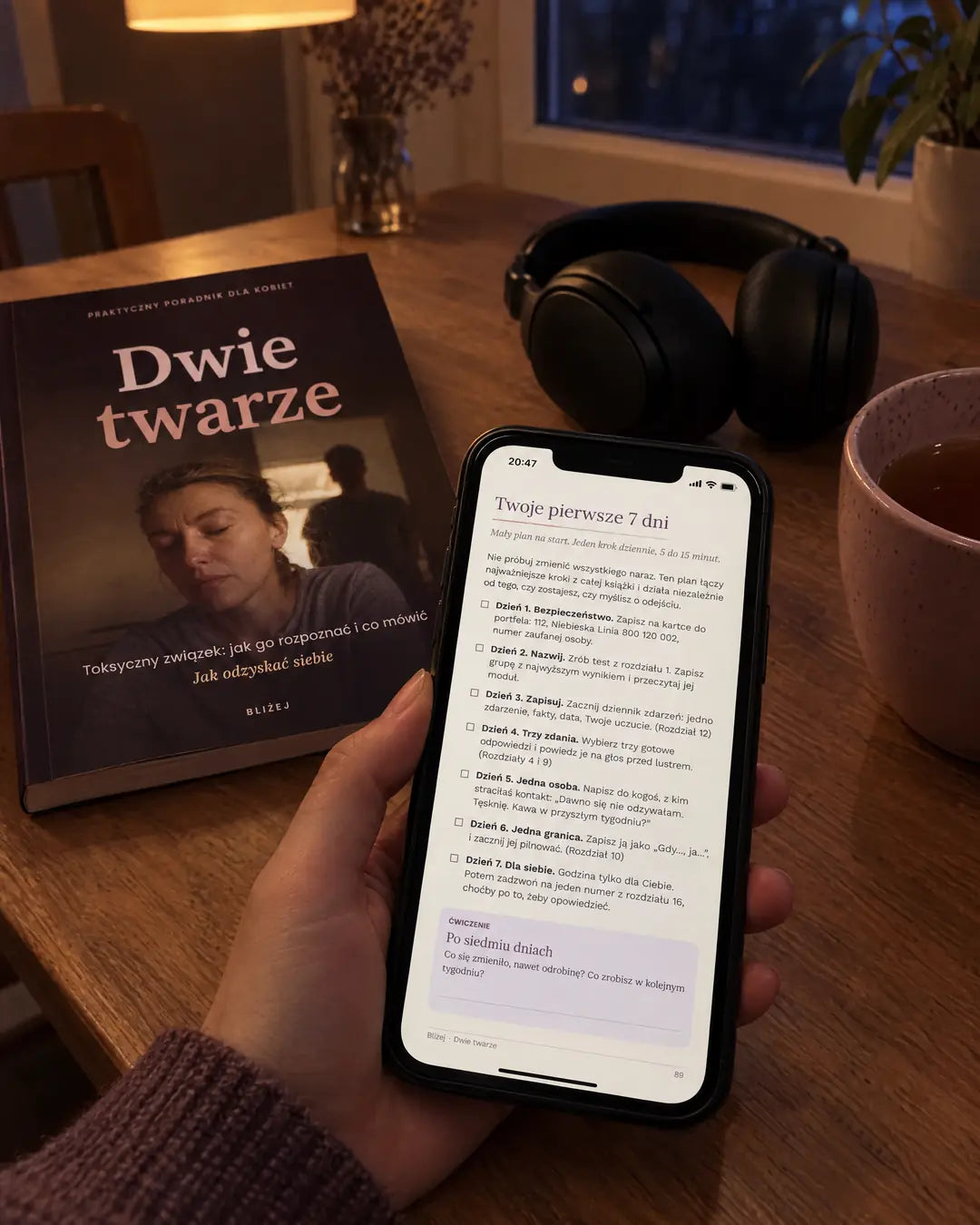 E-book „Dwie twarze” jako PDF w telefonie, obok wydruk okładki, słuchawki i herbata na stole wieczorem